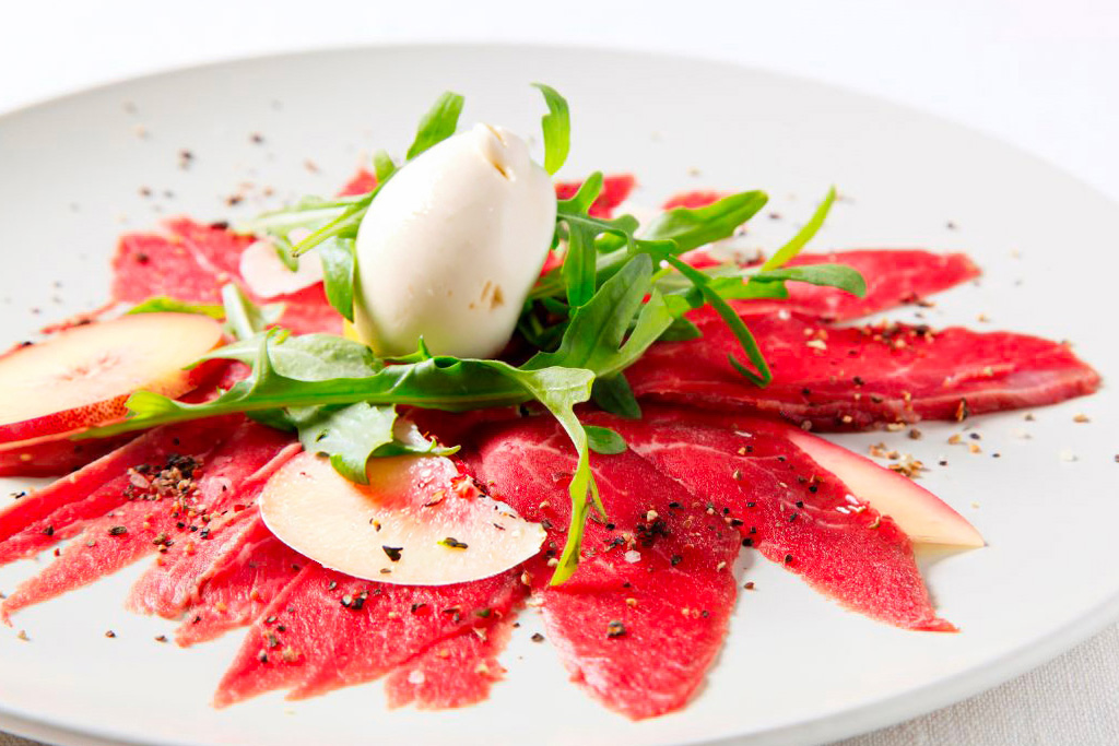 Un carpaccio de boeuf avec des tranches de pêches et une burrata dans une assiette blanche