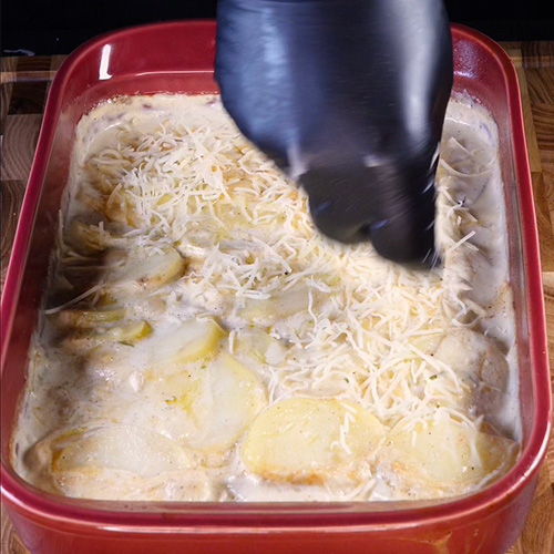 Une main gantée noire qui ajoute du fromage râpé sur un gratin dauphinois