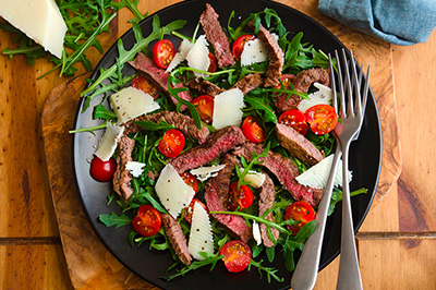 Une salade composée de tranches de bavette de boeuf, tomates, copeaux de parmesan et roquette servie dans une assiette noire